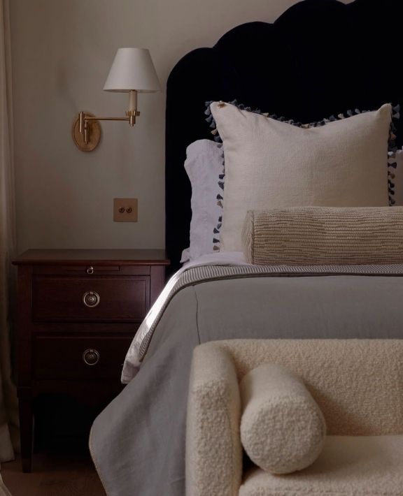 A cozy bedroom corner with a dark headboard, white pillows, a beige bolster, a gray blanket, a wooden nightstand with drawers, a wall-mounted lamp, and a cream-colored boucle chair in the foreground.