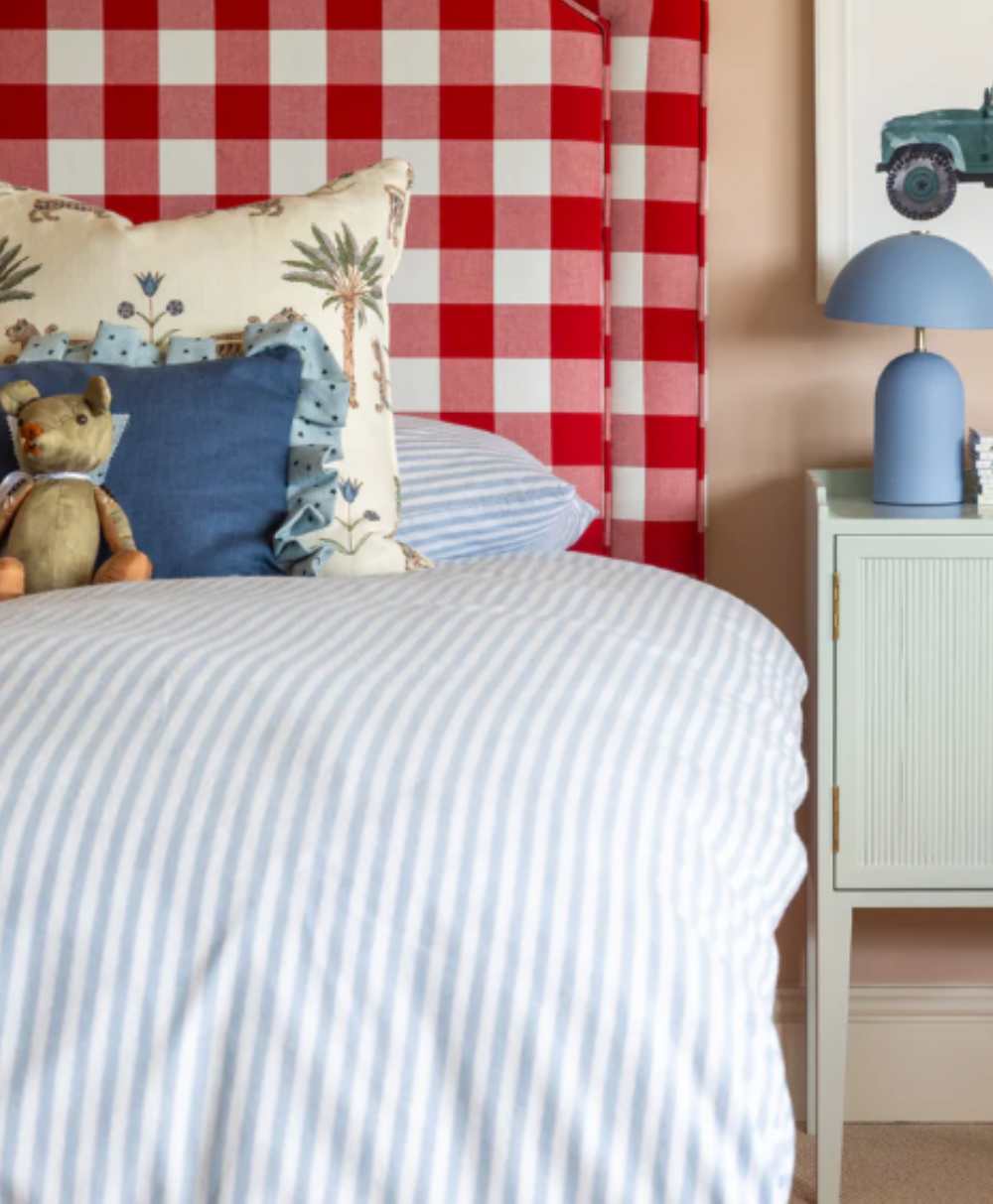 A cozy bed with blue and white striped bedding, a teddy bear, and patterned pillows sits next to a pale green nightstand with a blue lamp and a toy truck decoration on the wall above. The headboard is red and white plaid.
