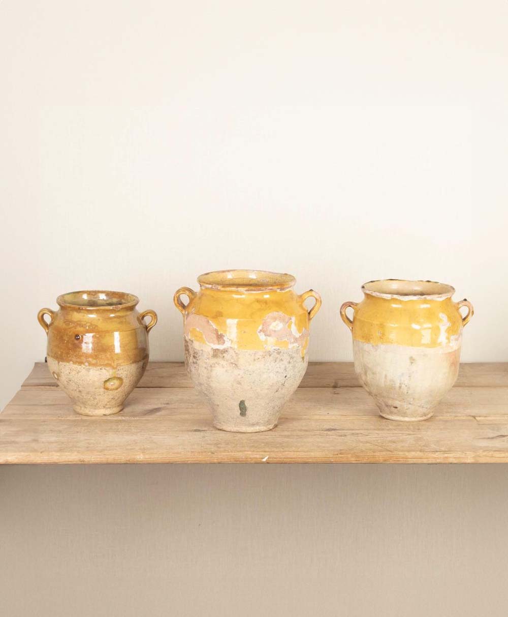 Three rustic, vintage clay pots with yellow glazed tops and handles are displayed on a wooden shelf against a plain, light-colored wall. The pots vary slightly in size and shape.