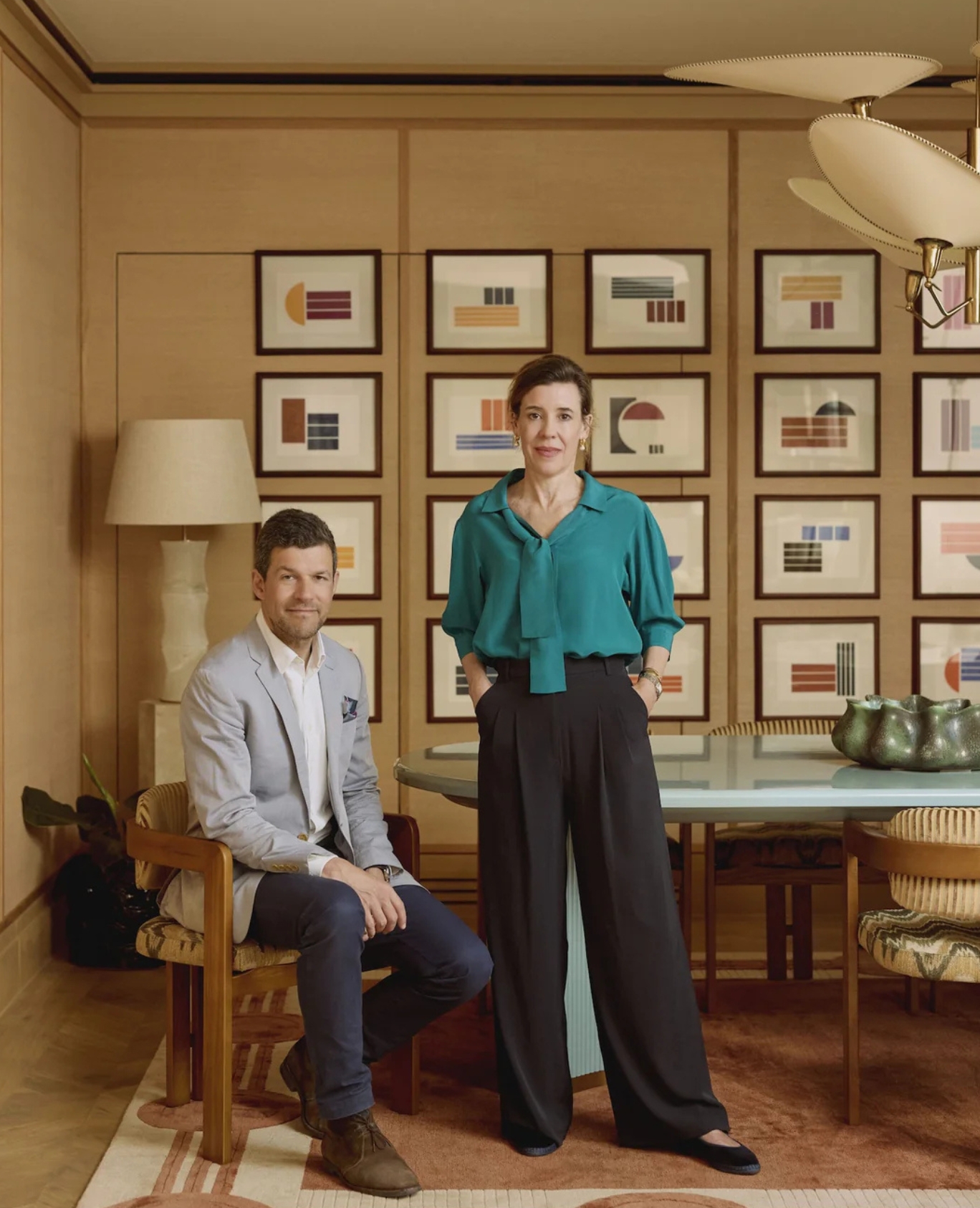 A man sits on a chair and a woman stands beside him in a stylish room with framed artwork on the wall, a large lamp, a table, and decorative objects. Both are dressed in smart, modern clothing.