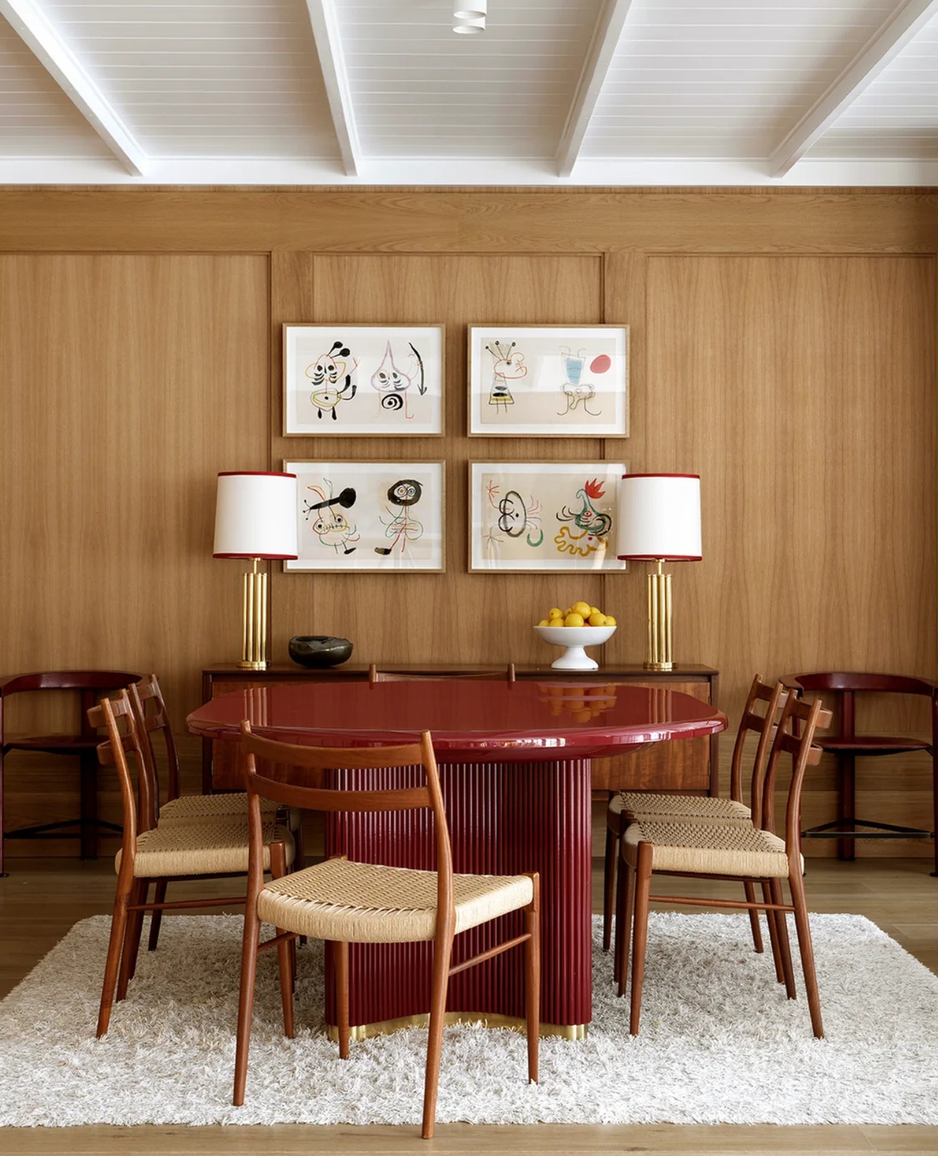 A modern dining room features a glossy red table, six wooden chairs with woven seats, two table lamps with white shades, and four framed artworks on a wood-paneled wall. A white rug covers the floor.