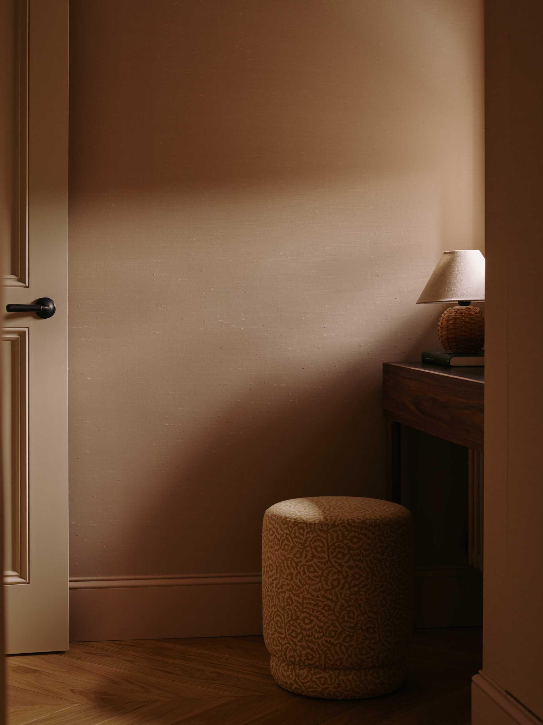 A softly lit corner of a room with beige walls, a closed white door, a small round upholstered stool, and a dark wooden table topped with a small lamp, casting a warm, cozy glow.