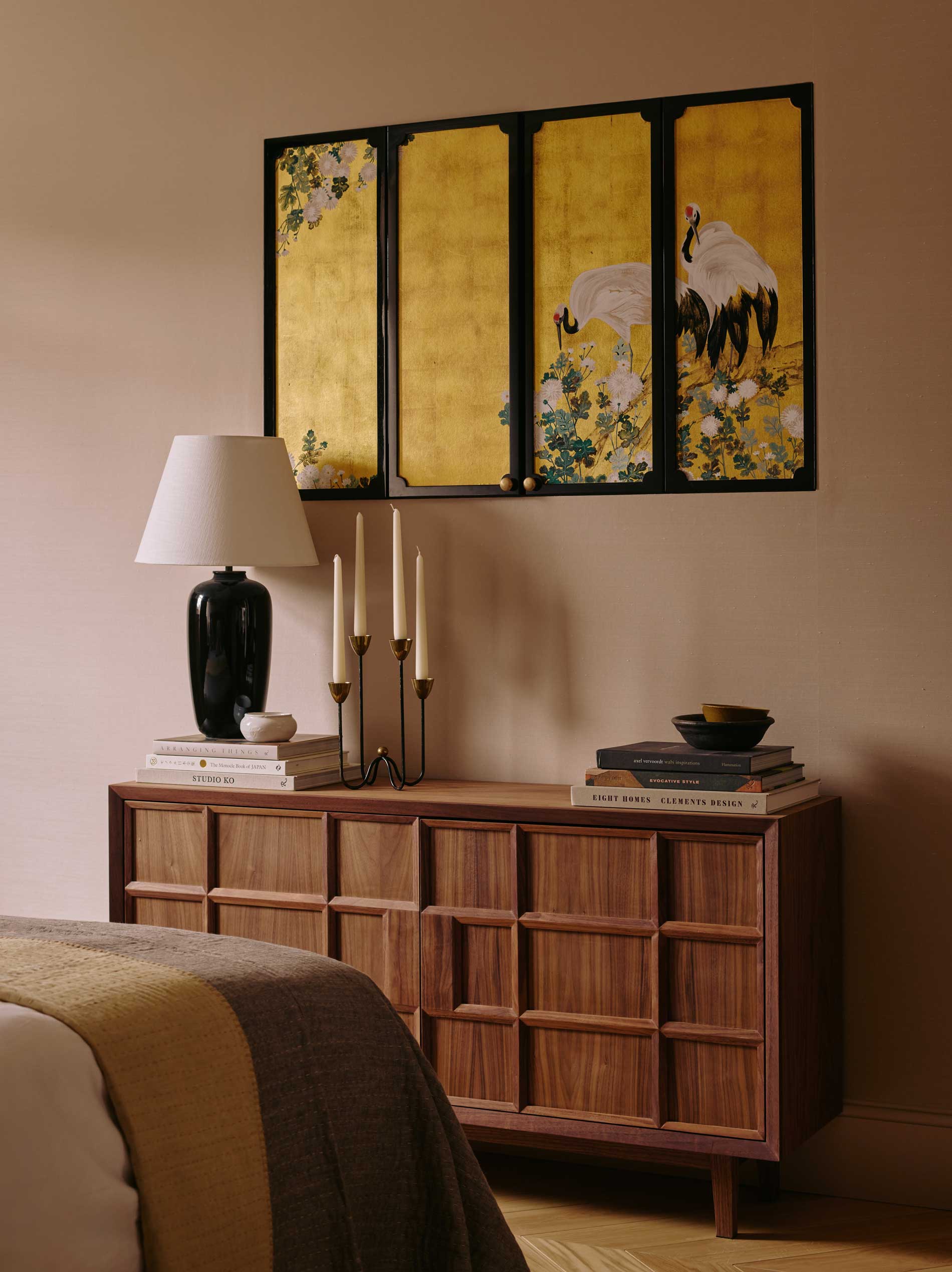 A wooden sideboard with books, a black lamp, candlesticks, and a bowl sits against a beige wall. Above it hangs a yellow and black artwork featuring cranes and flowers. Part of a bed with a blanket is visible in the foreground.