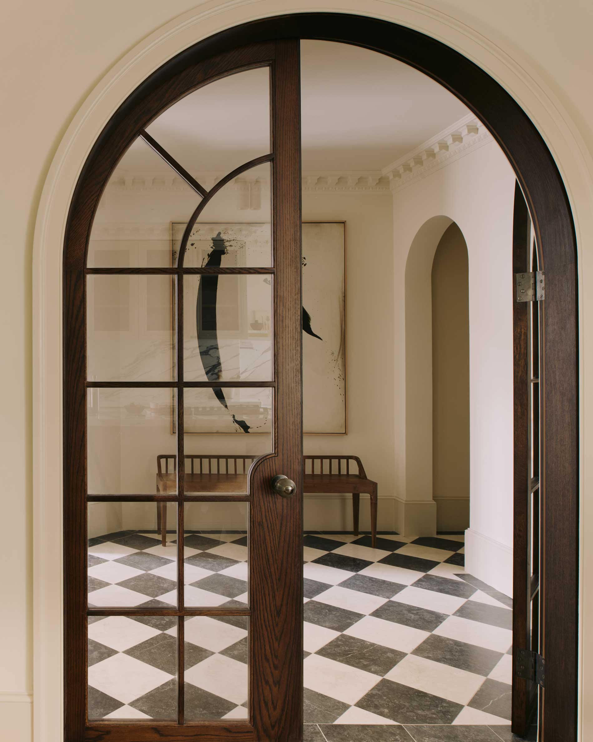 An arched wooden door with glass panes opens to a hallway with black-and-white checkered floor tiles, a wooden bench, and abstract art on the wall. The space has soft, neutral tones and elegant architectural details.