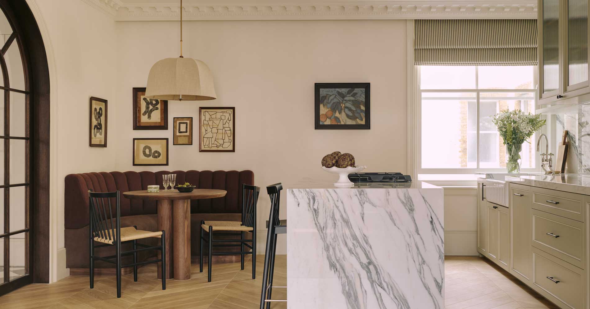 A stylish kitchen and dining area with a marble island, a round wooden table with dark chairs, a brown cushioned bench, wall art, large windows, and light cabinets with flowers in a vase.