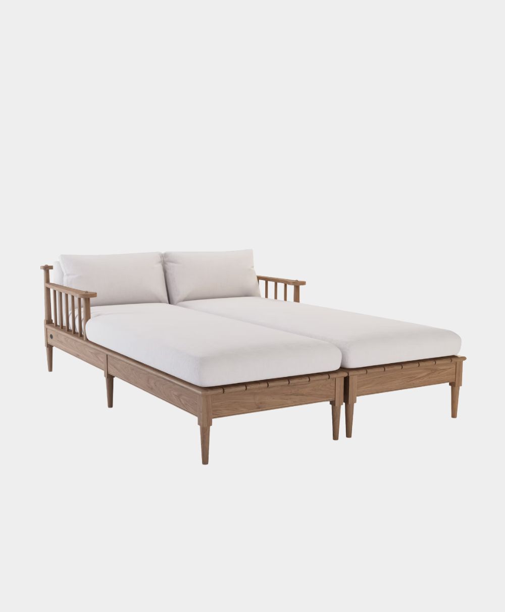 A modern wooden daybed with light wood frame, spindled armrests, angled legs, and white cushions on the back and seat, designed for two people, set against a plain white background.