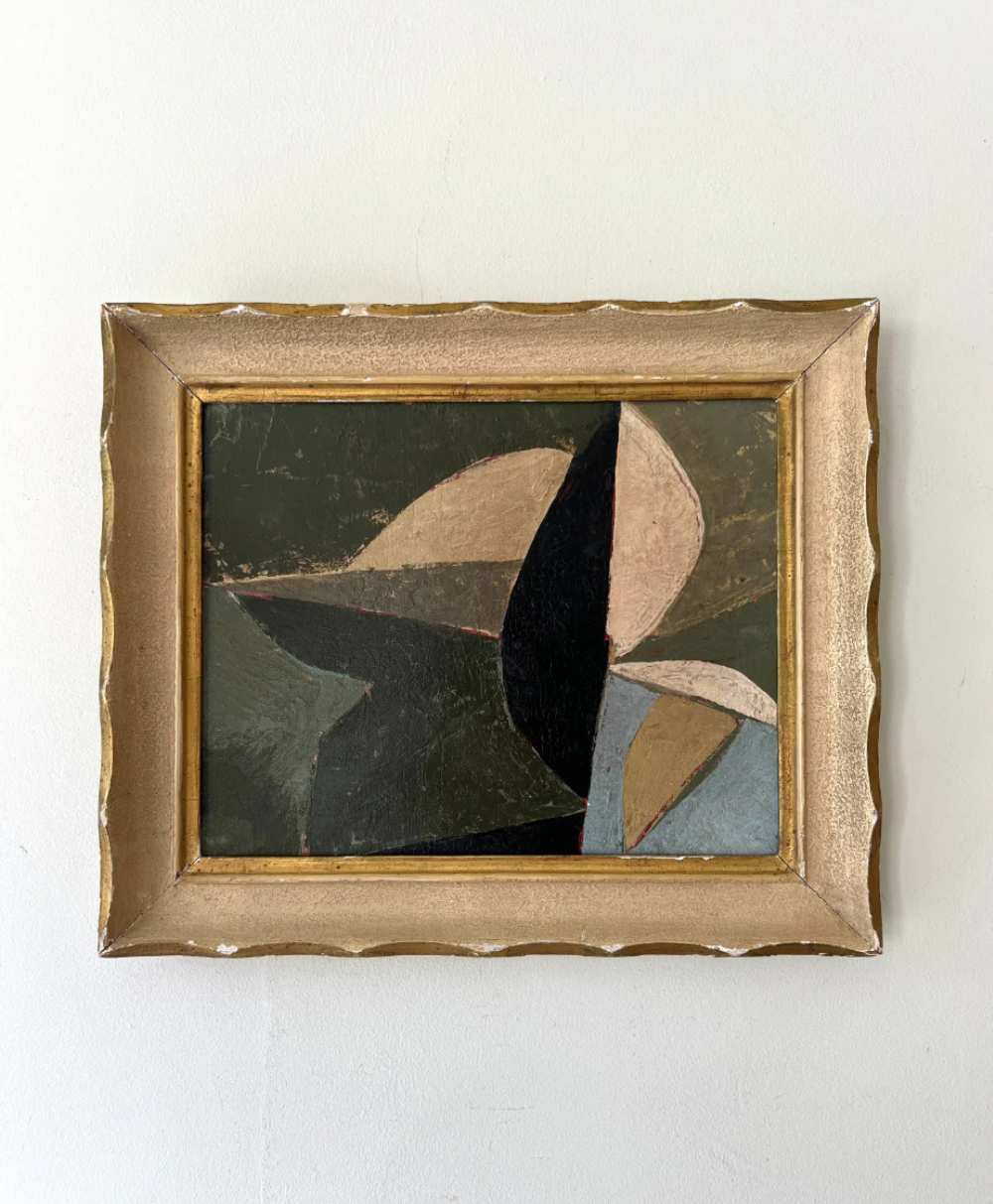 A framed abstract painting with overlapping geometric shapes in muted tones of beige, black, olive green, and blue, set against a plain light-colored wall. The frame is gold with a wavy design.
