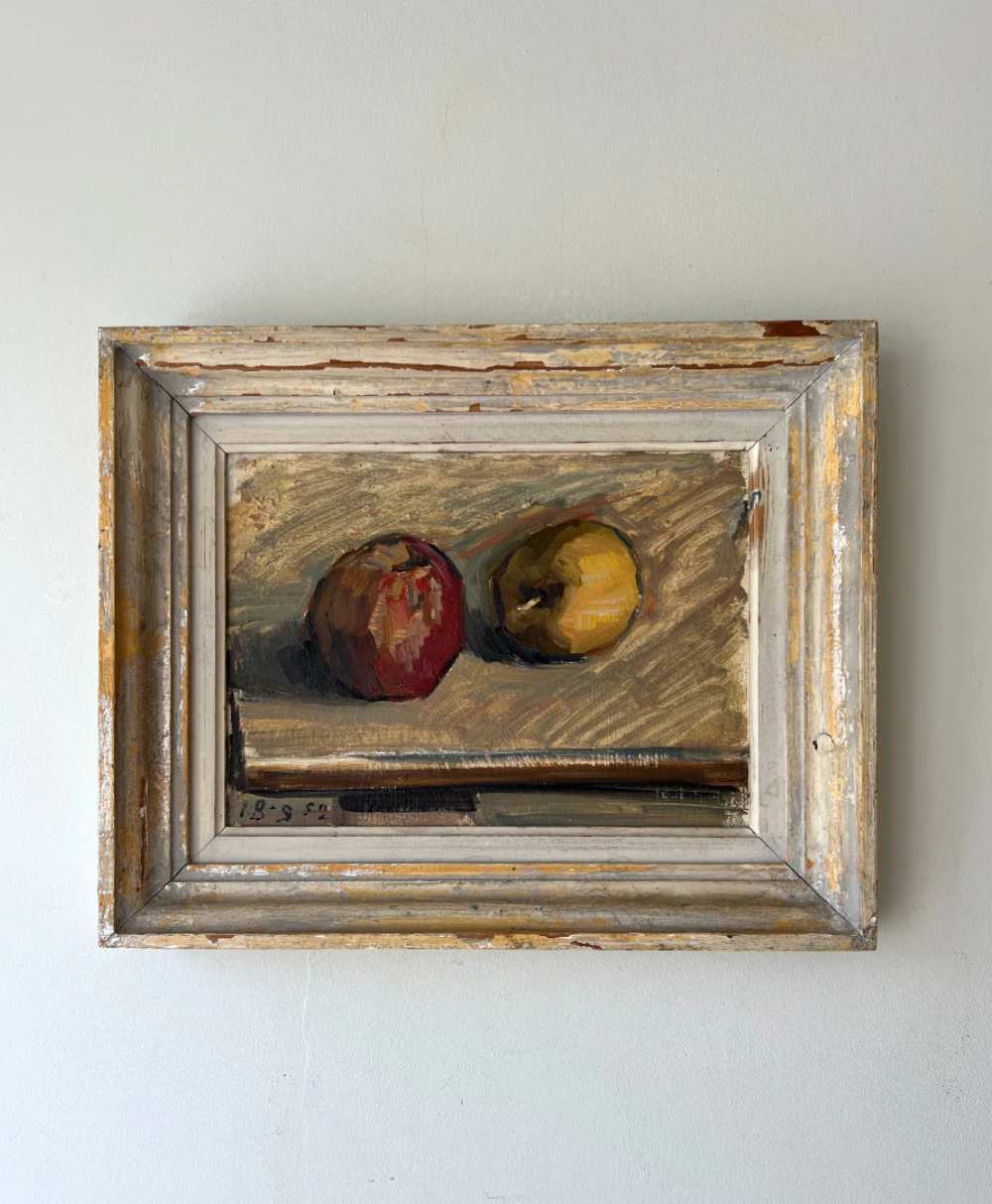 A framed painting of two apples, one red and one yellow, resting on a flat surface. The frame is rustic, with chipped paint and a weathered appearance, hanging on a plain light-colored wall.
