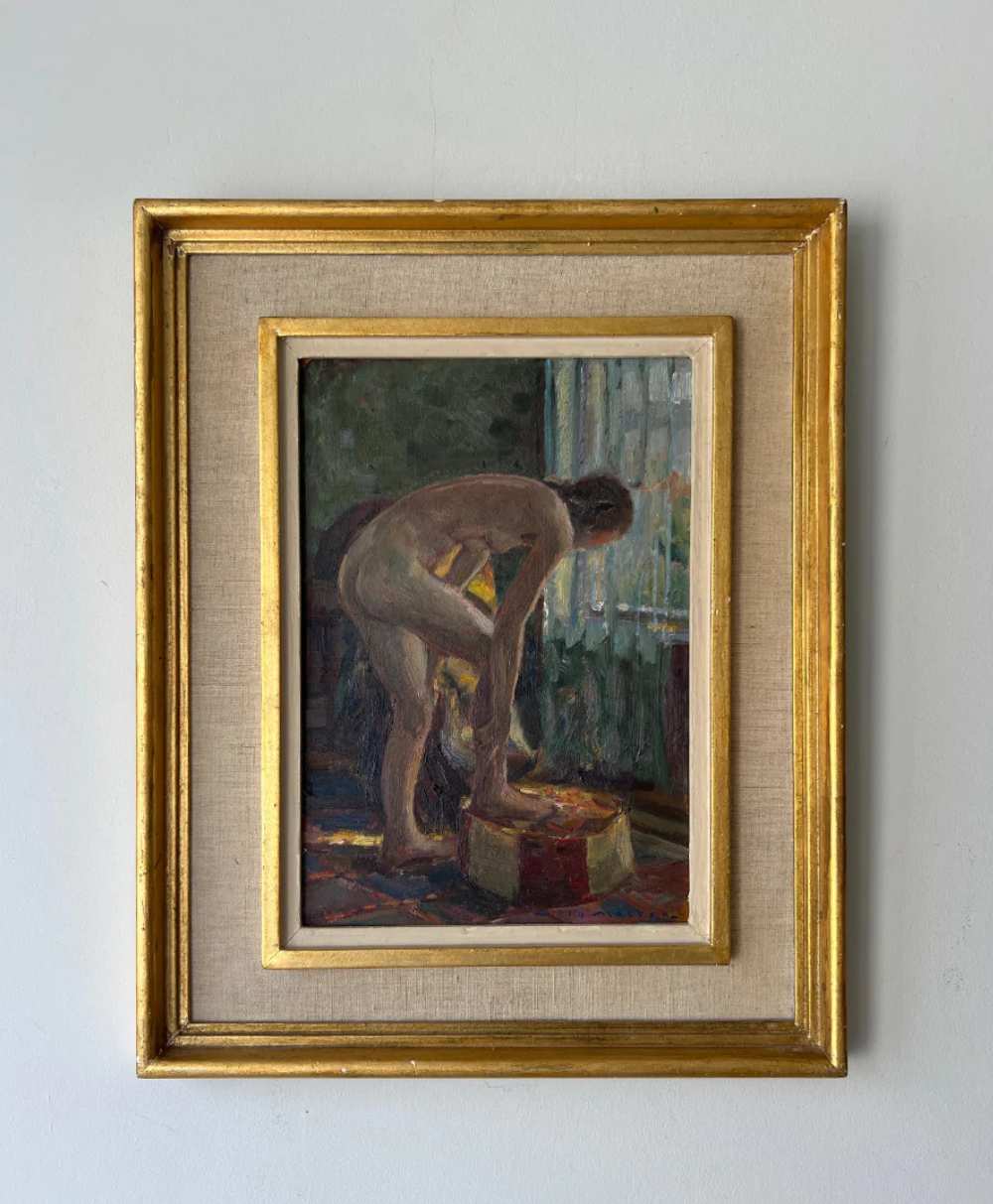 A framed painting of a nude person bending over in a dimly lit room, with light streaming through a window. The frame has a gold outer border and a beige linen mat surrounding the artwork.
