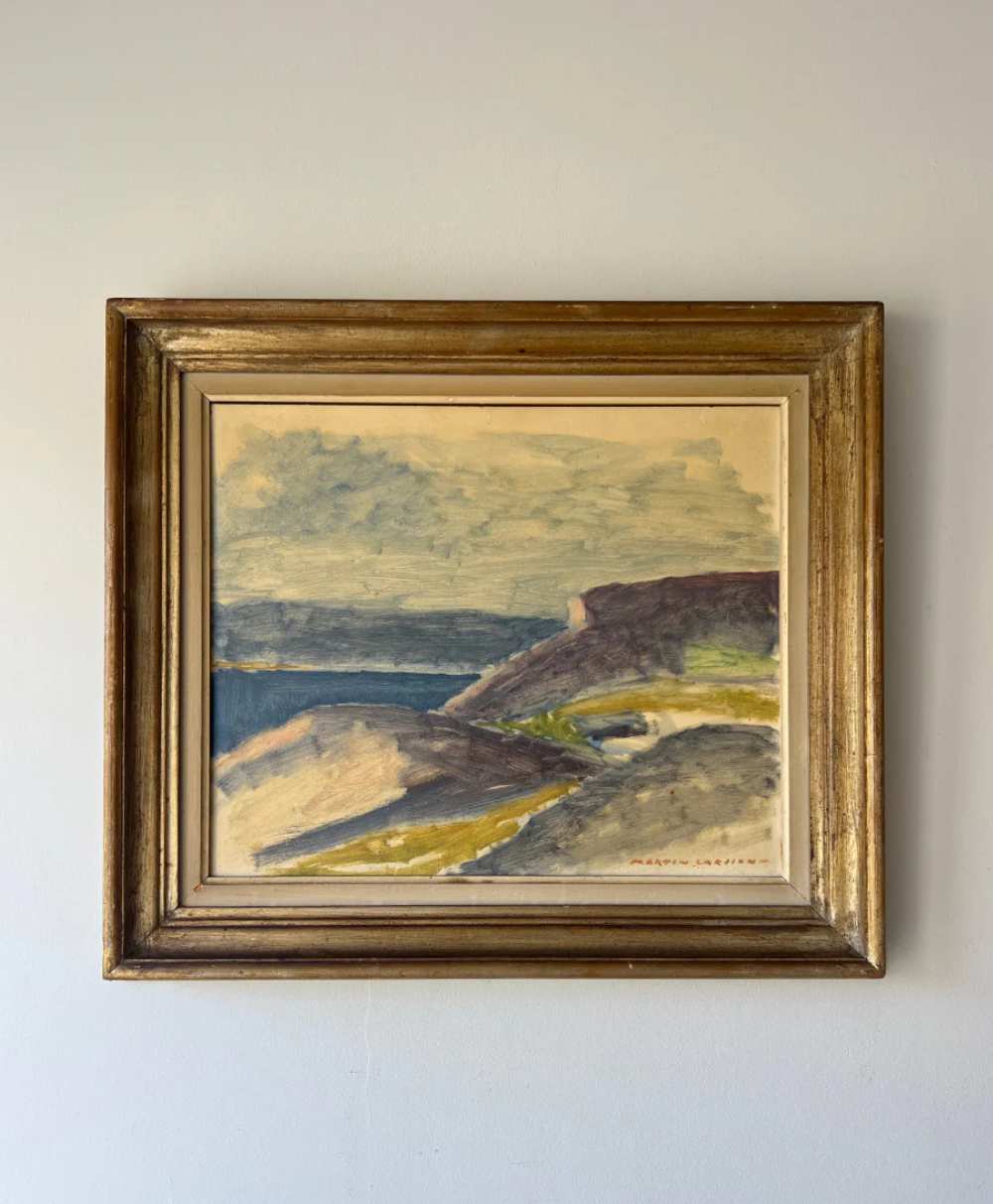 A framed landscape painting featuring a coastal scene with cliffs, water, and a cloudy sky, displayed on a plain wall.