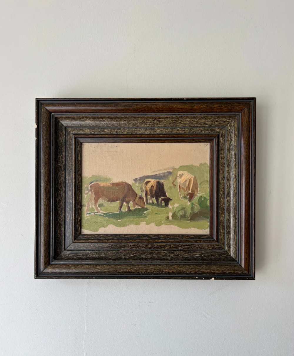 A small framed painting of three cows grazing in a grassy field, set against a light-colored wall. The painting uses soft, muted colors and is bordered by a wide, dark wooden frame.
