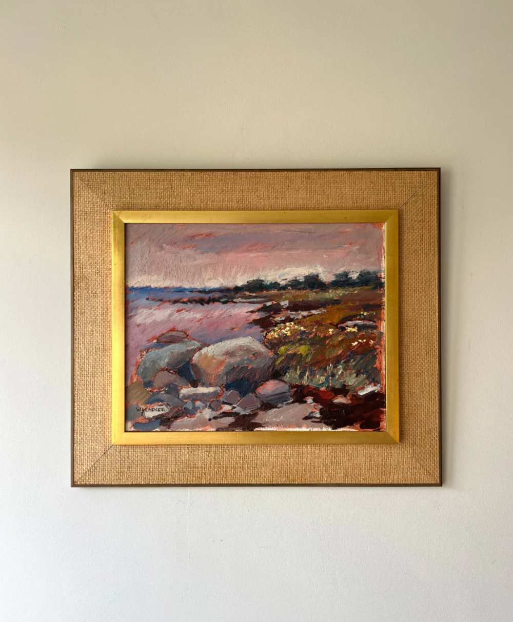 Framed landscape painting featuring a rocky shoreline with greenery, a calm body of water, and distant trees under a pink and purple sky, hung on a plain light-colored wall.