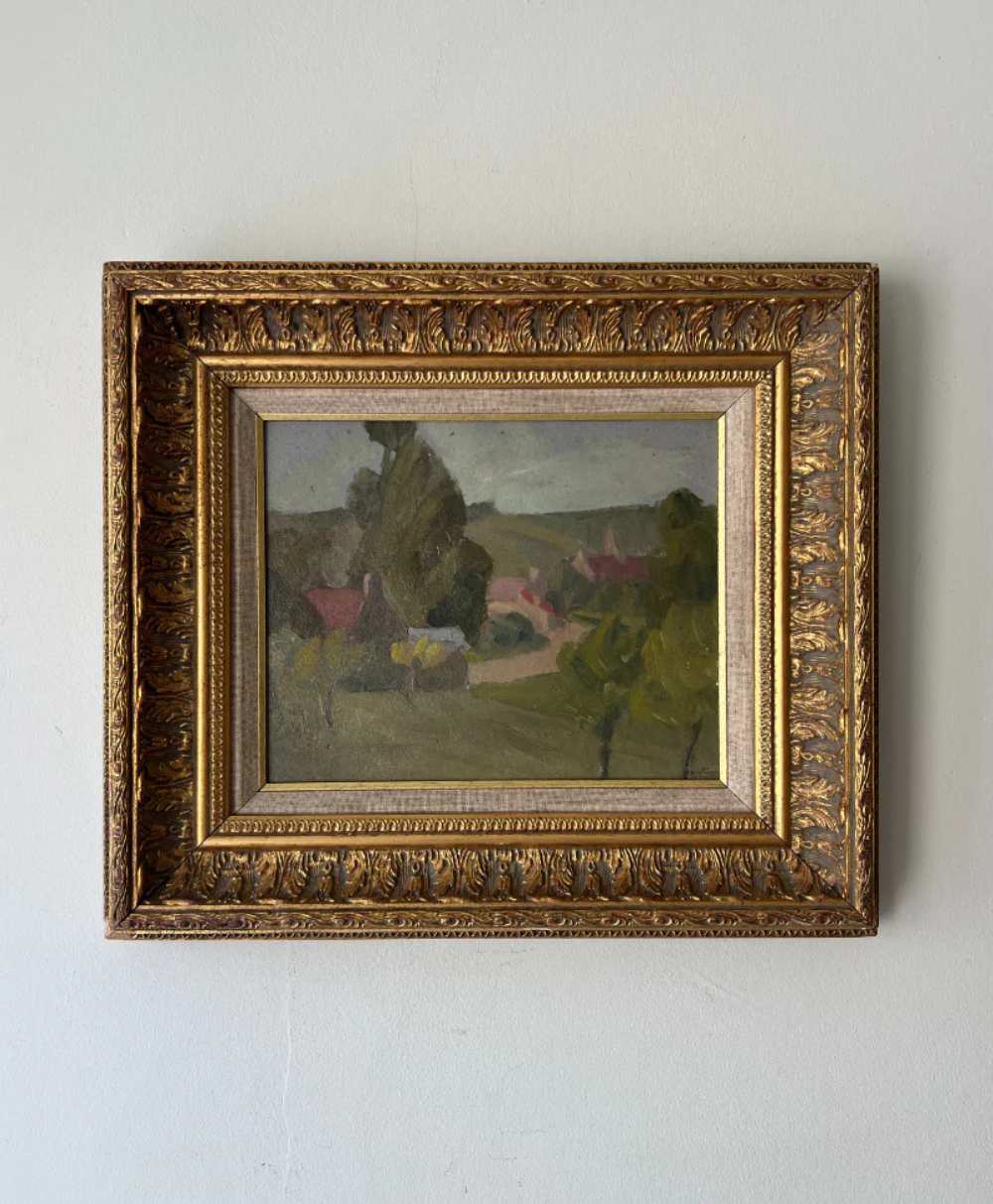 A framed landscape painting with muted colors, featuring trees, hills, and a few buildings. The painting is enclosed in an ornate, gold-toned frame and hung on a plain, light-colored wall.