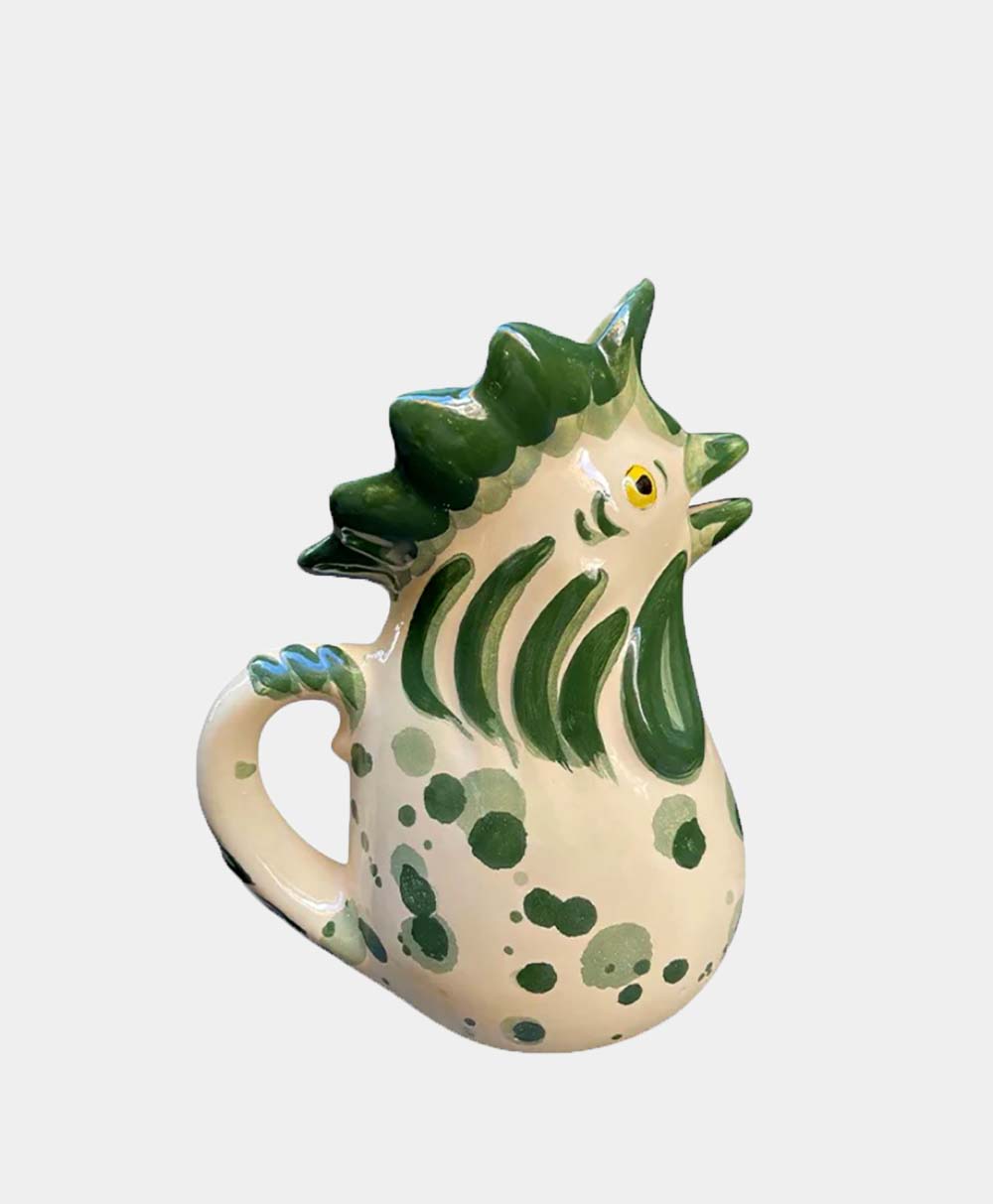 A ceramic pitcher shaped like a roosters head, painted white with green spots and accents, featuring a handle and an open beak spout.