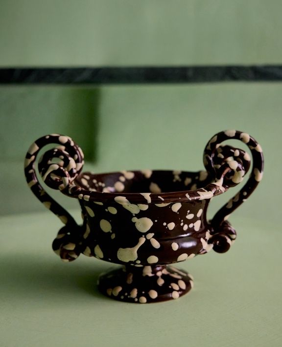A decorative, dark brown urn with two ornate handles and cream-colored spots sits on a light green surface against a matching green background.