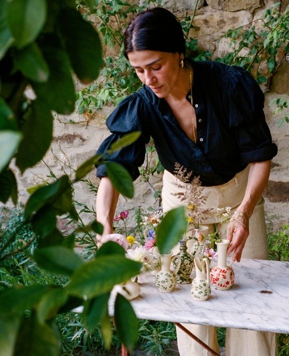 A woman with dark hair in a black blouse and light pants arranges flowers in small vases on a marble table outdoors, surrounded by green plants and a stone wall.