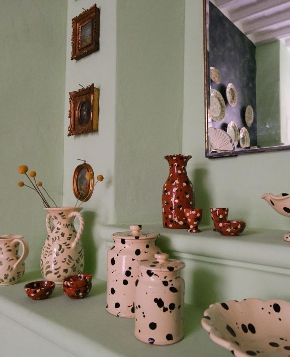 A collection of cream and rust-colored pottery with black spots displayed on green shelves, alongside small framed pictures on the wall and a mirror reflecting more pottery.
