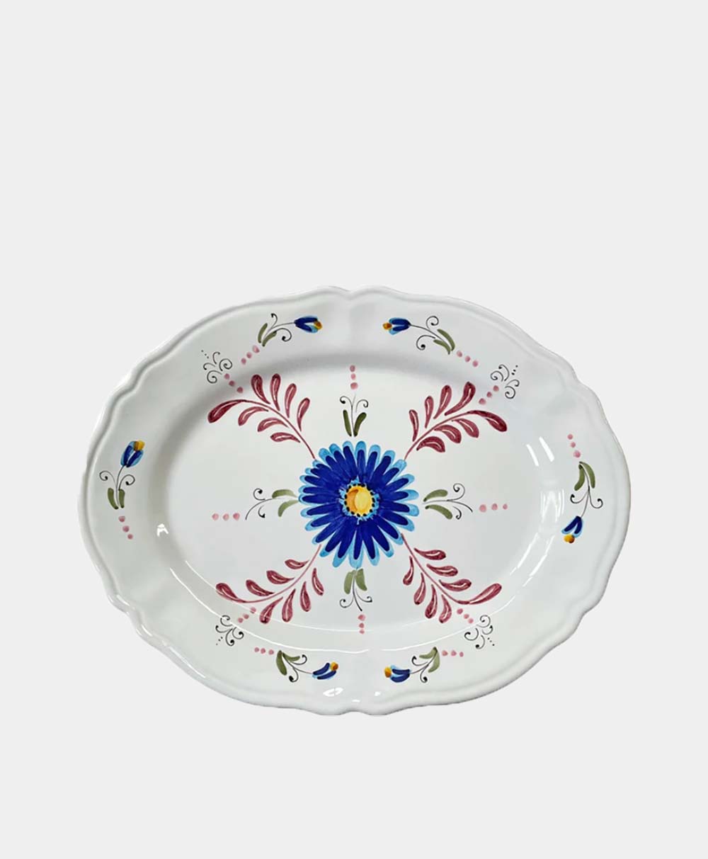 An oval ceramic plate with scalloped edges, featuring a hand-painted blue flower in the center, surrounded by red, yellow, green, and blue decorative floral designs on a white background.