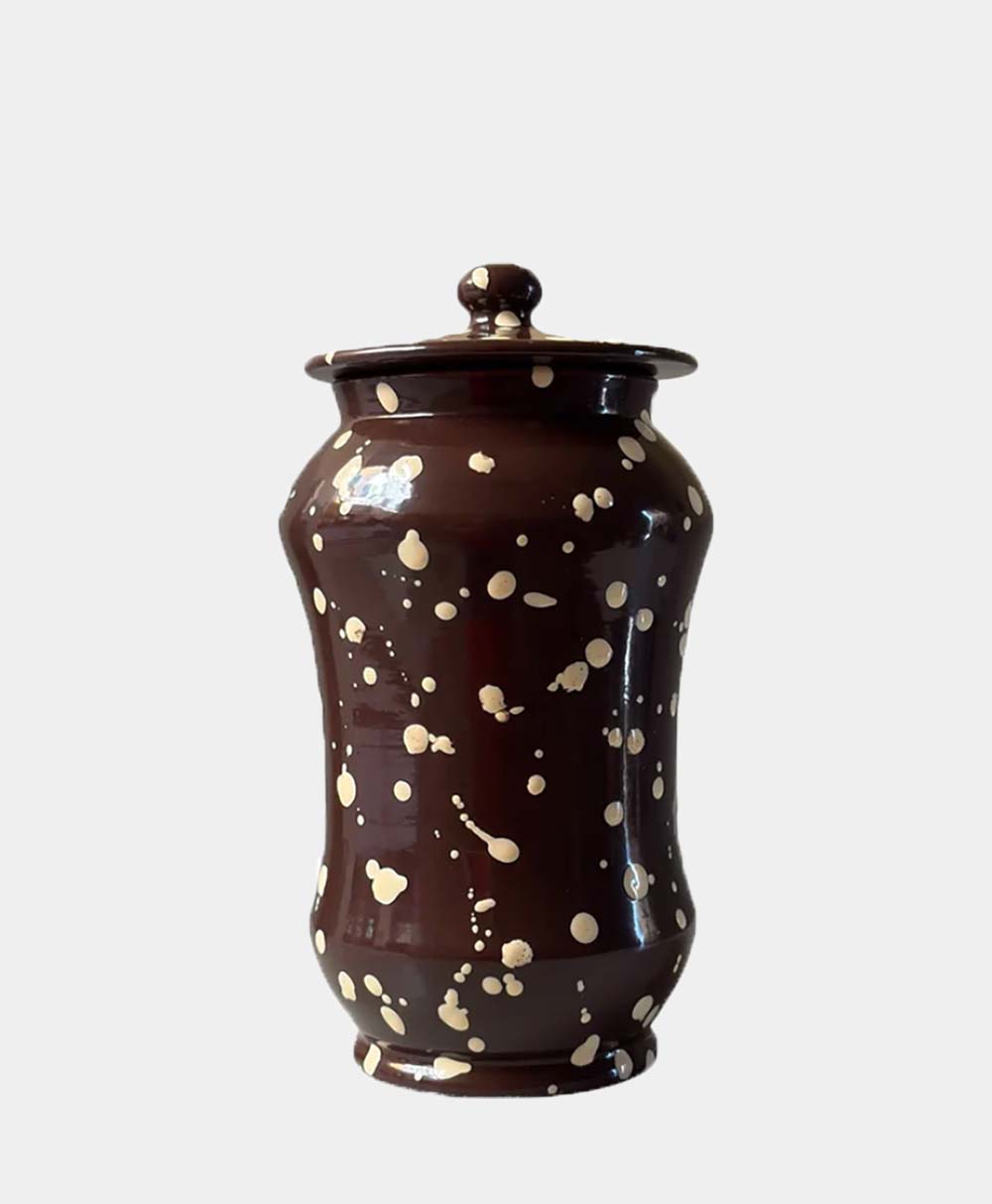 A tall, brown ceramic jar with a matching lid, decorated with irregular cream-colored splatter patterns, set against a plain light background.