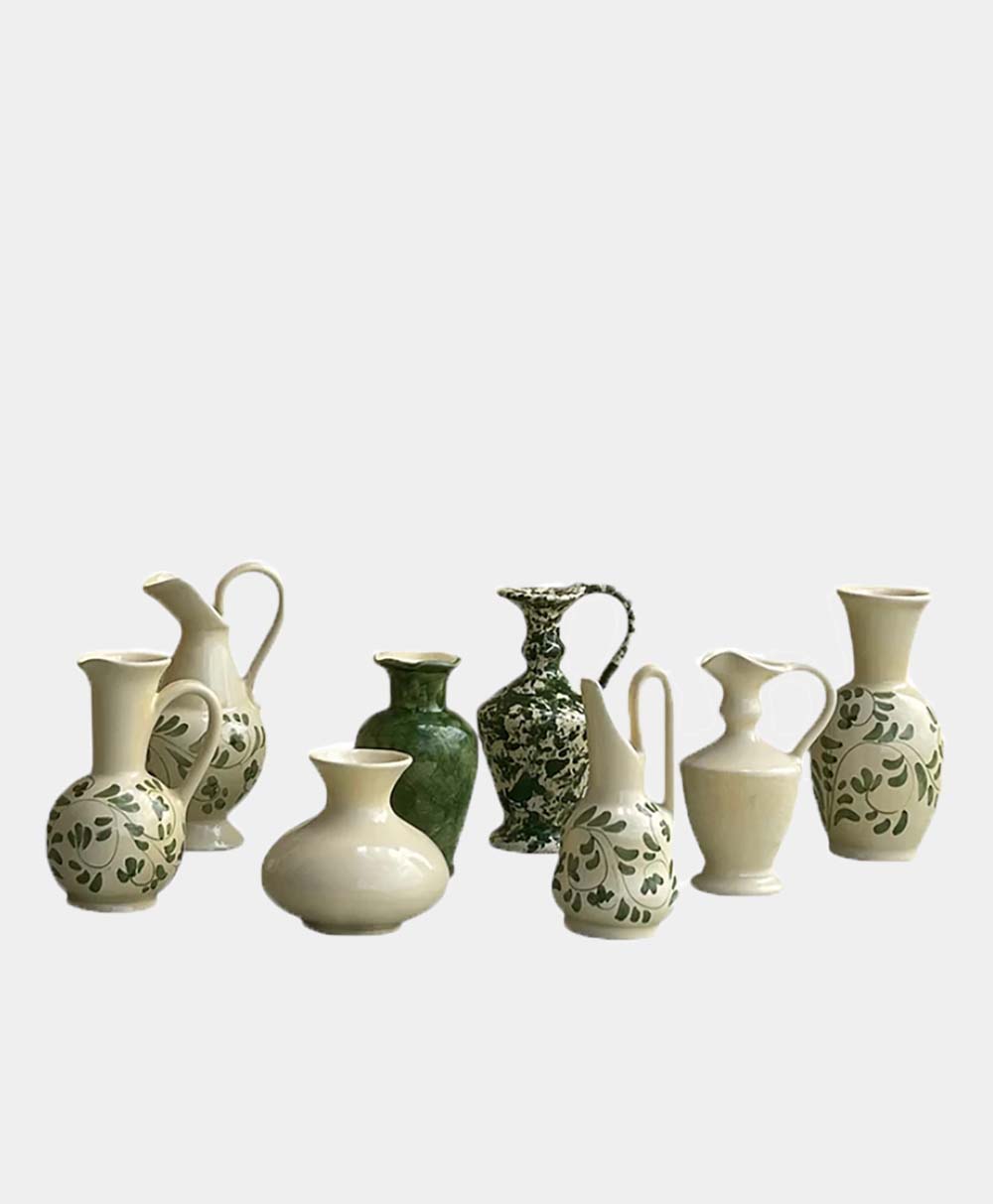 A group of eight ceramic vases in various shapes and sizes, featuring white, green, and floral patterns, are arranged together against a plain light gray background.