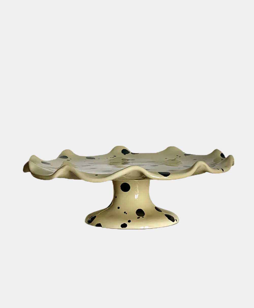 A ceramic cake stand with a wavy, ruffled edge and a wide base. The stand is cream-colored with irregular black spots, giving it a playful, modern look. The background is plain white.