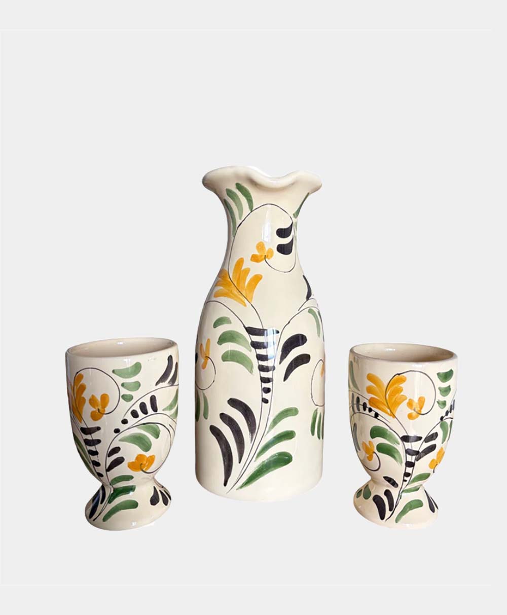 A ceramic set with a tall pitcher and two matching cups, all decorated with hand-painted yellow flowers, green leaves, and black accents on a cream background.