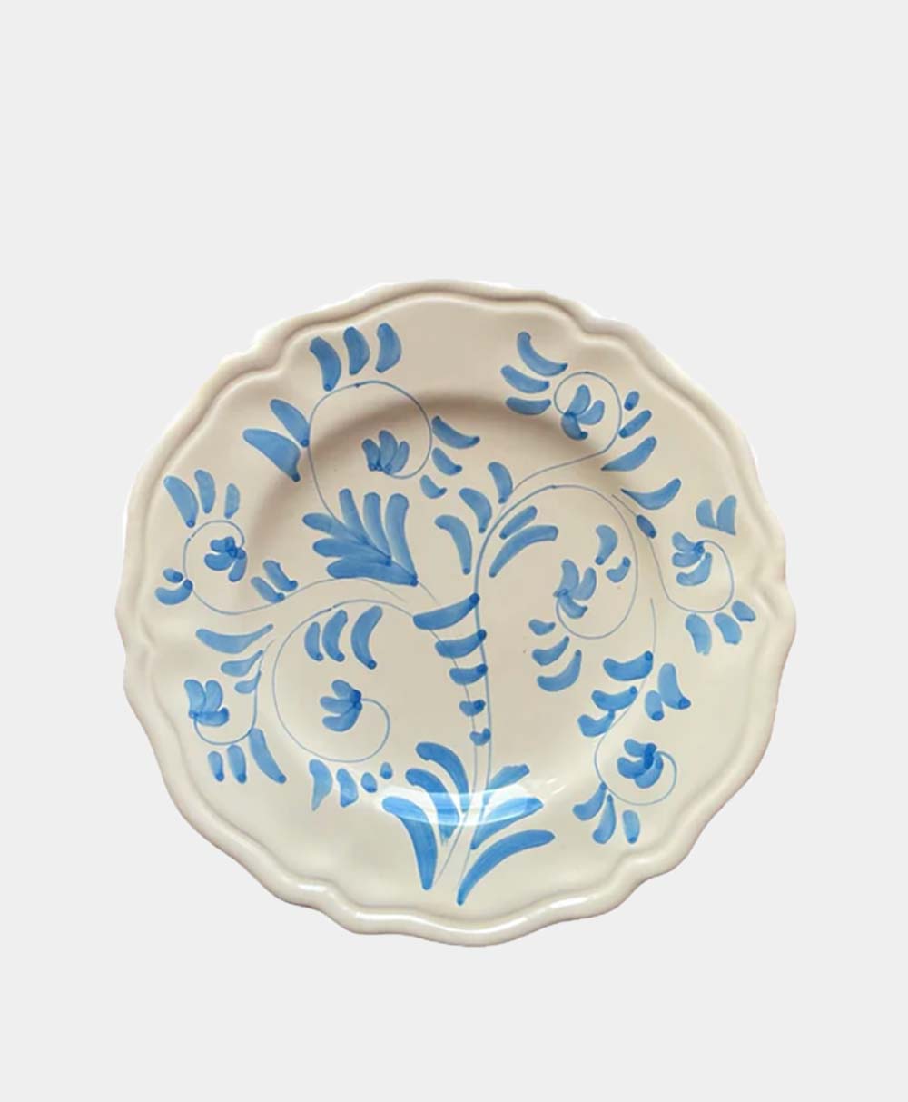 A round, cream-colored ceramic plate with a scalloped edge, decorated with a hand-painted blue floral and leaf design. The background is plain and light grey.