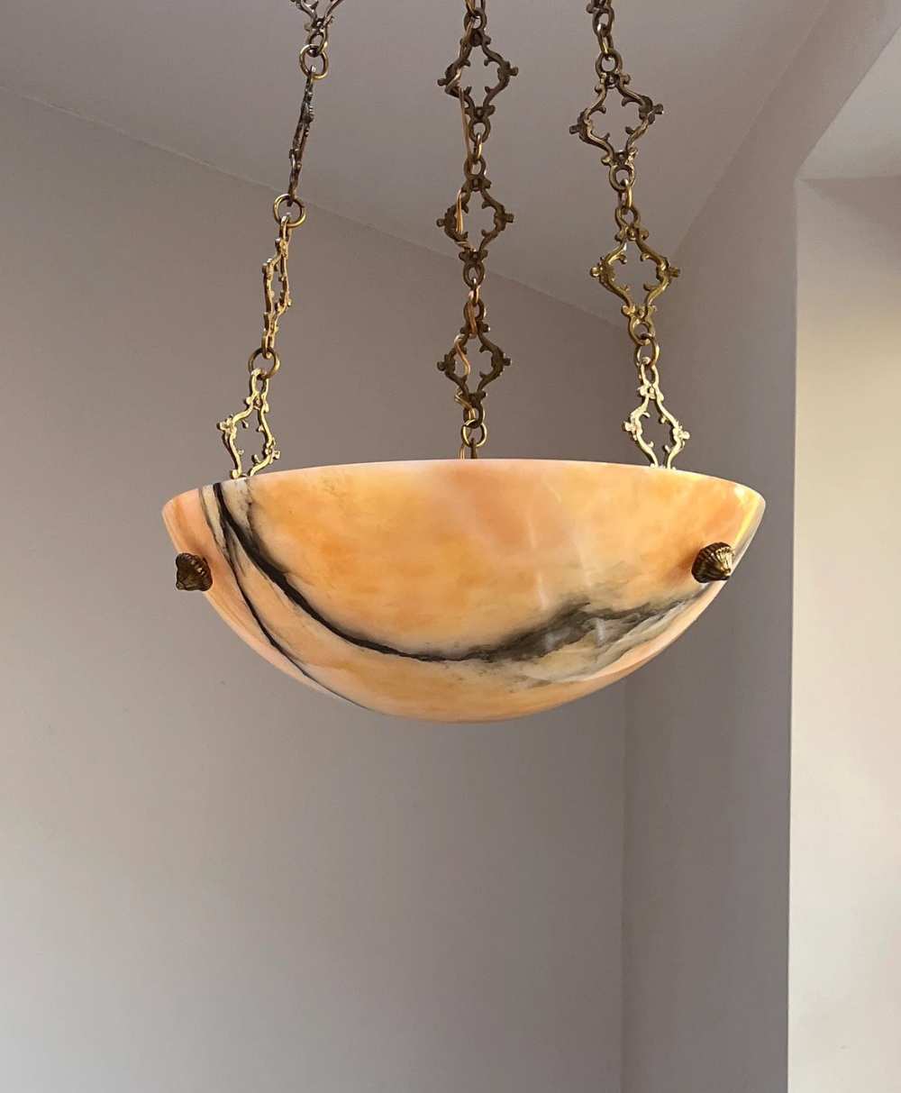 A marble-patterned, bowl-shaped pendant light with warm orange and brown tones hangs from the ceiling by ornate gold chains against a neutral-colored wall.