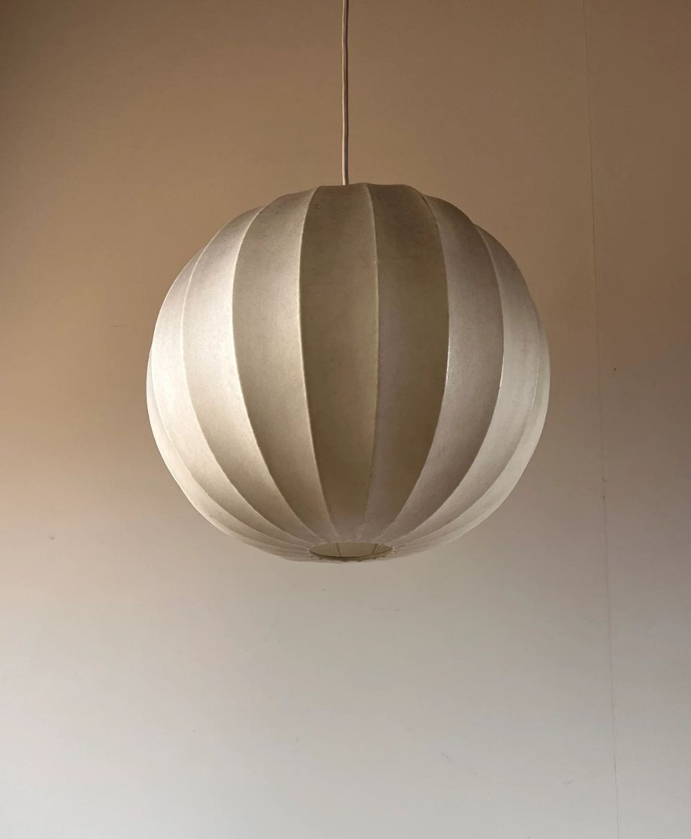 A spherical, white paper lantern lamp with vertical ridges hangs from the ceiling against a plain beige wall. The lamp is centered in the image and softly illuminated.