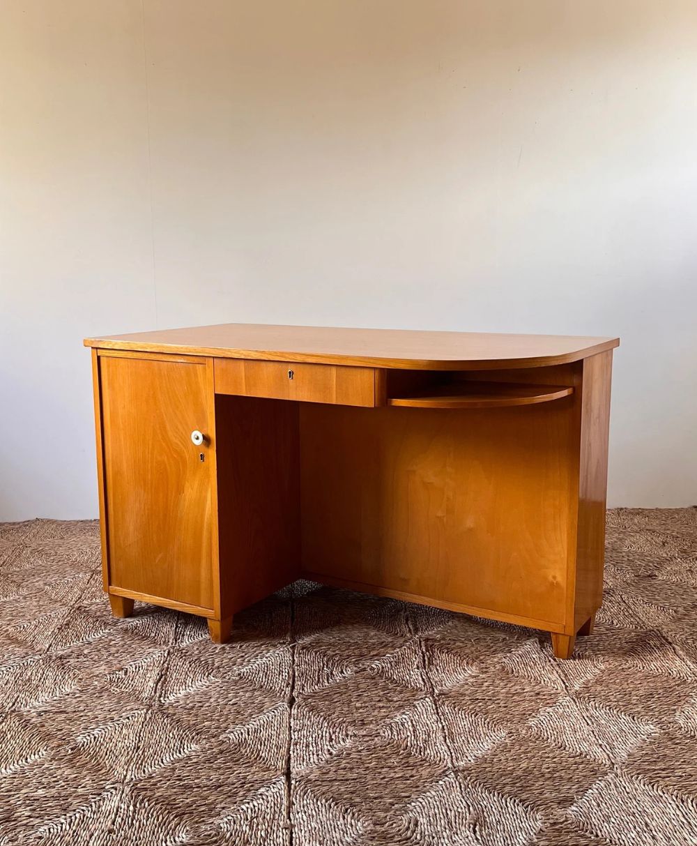 A vintage wooden desk with a curved front, a single drawer in the center, and a cabinet on the left side, standing on a patterned carpet against a plain light-colored wall.