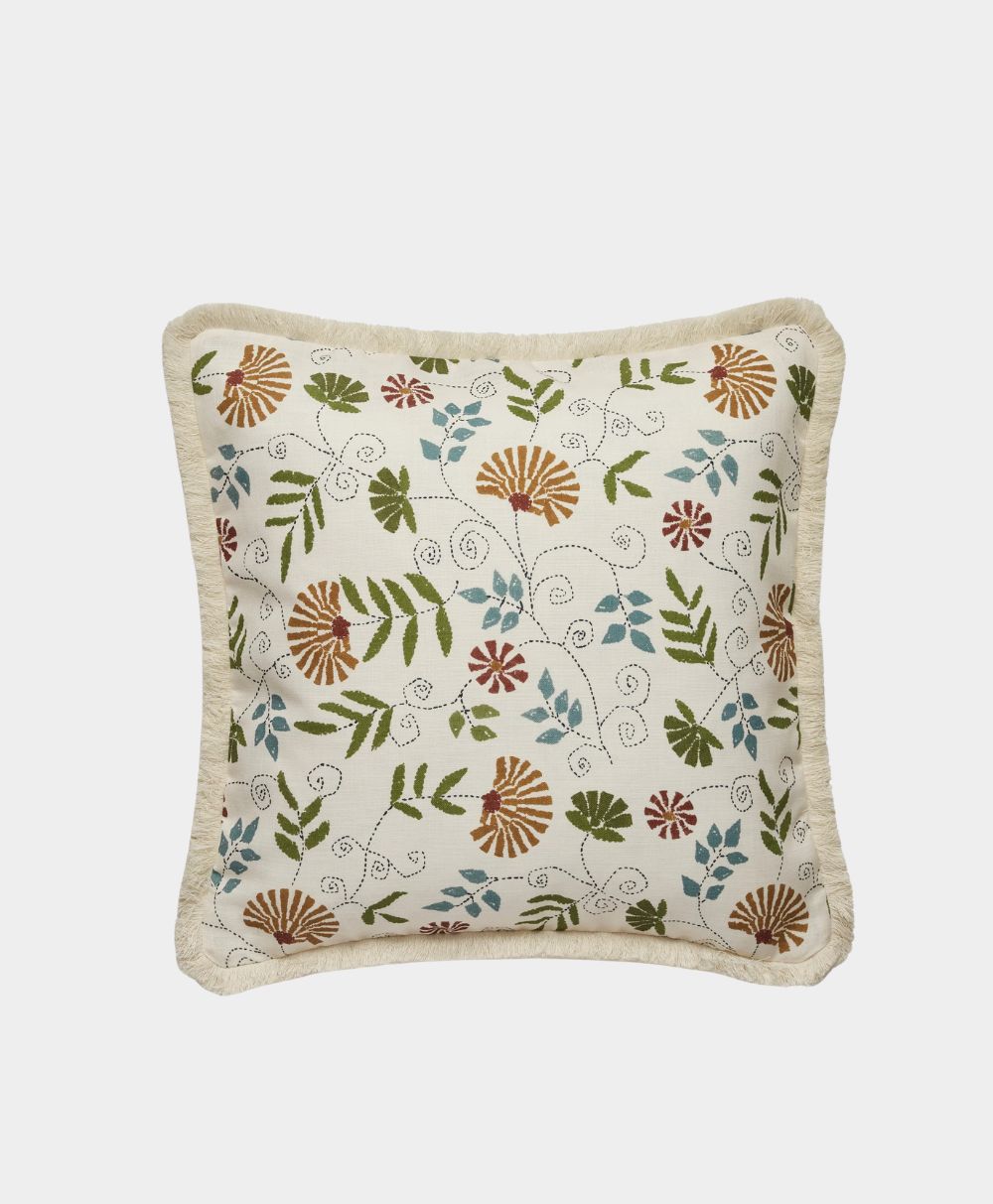 A square cream-colored pillow with a fringed edge features an embroidered floral pattern in green, brown, blue, and red on the front. The background is plain white.