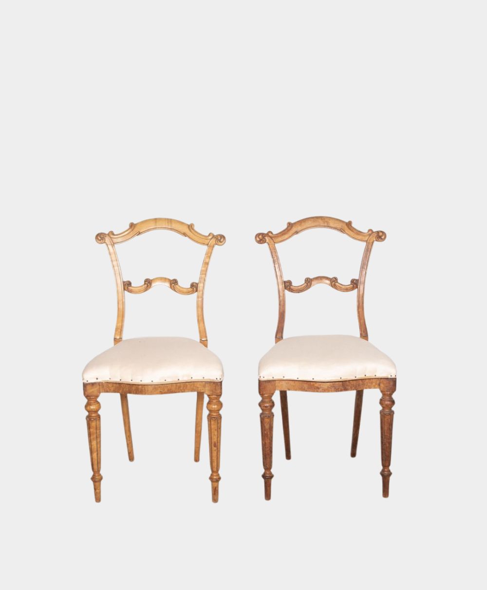 Two vintage wooden chairs with light upholstered seats, ornate carved backs, and spindle legs, set against a plain white background.