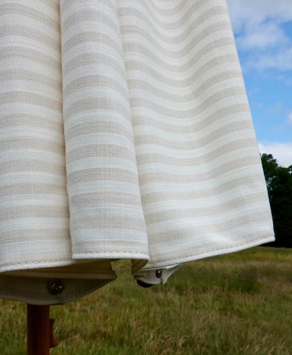 A close-up of a beige and white striped outdoor umbrella, partially open, with a grassy field and trees in the blurred background under a partly cloudy sky.