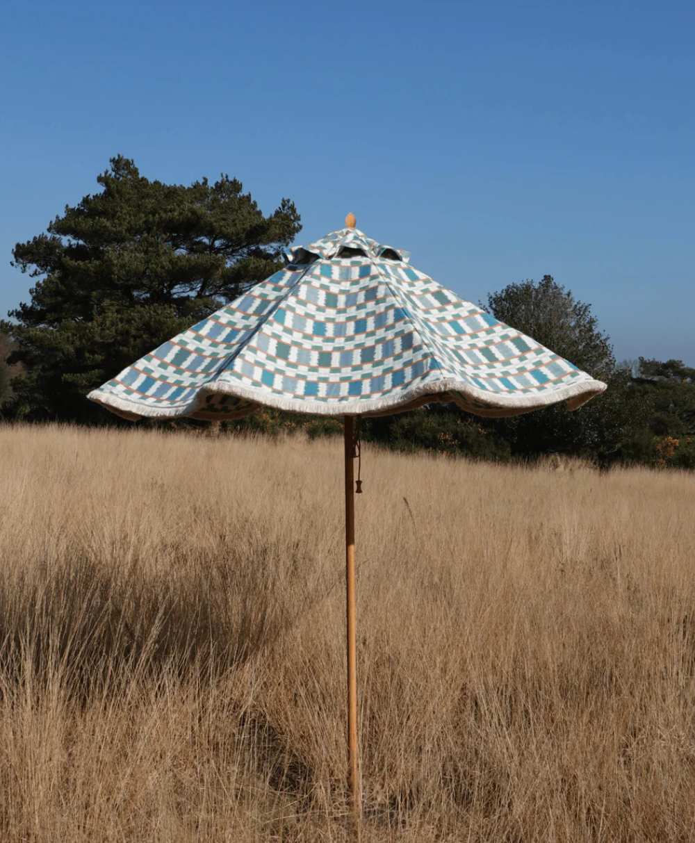 A blue and white checkered outdoor umbrella stands in a dry grassy field with trees and a clear blue sky in the background.