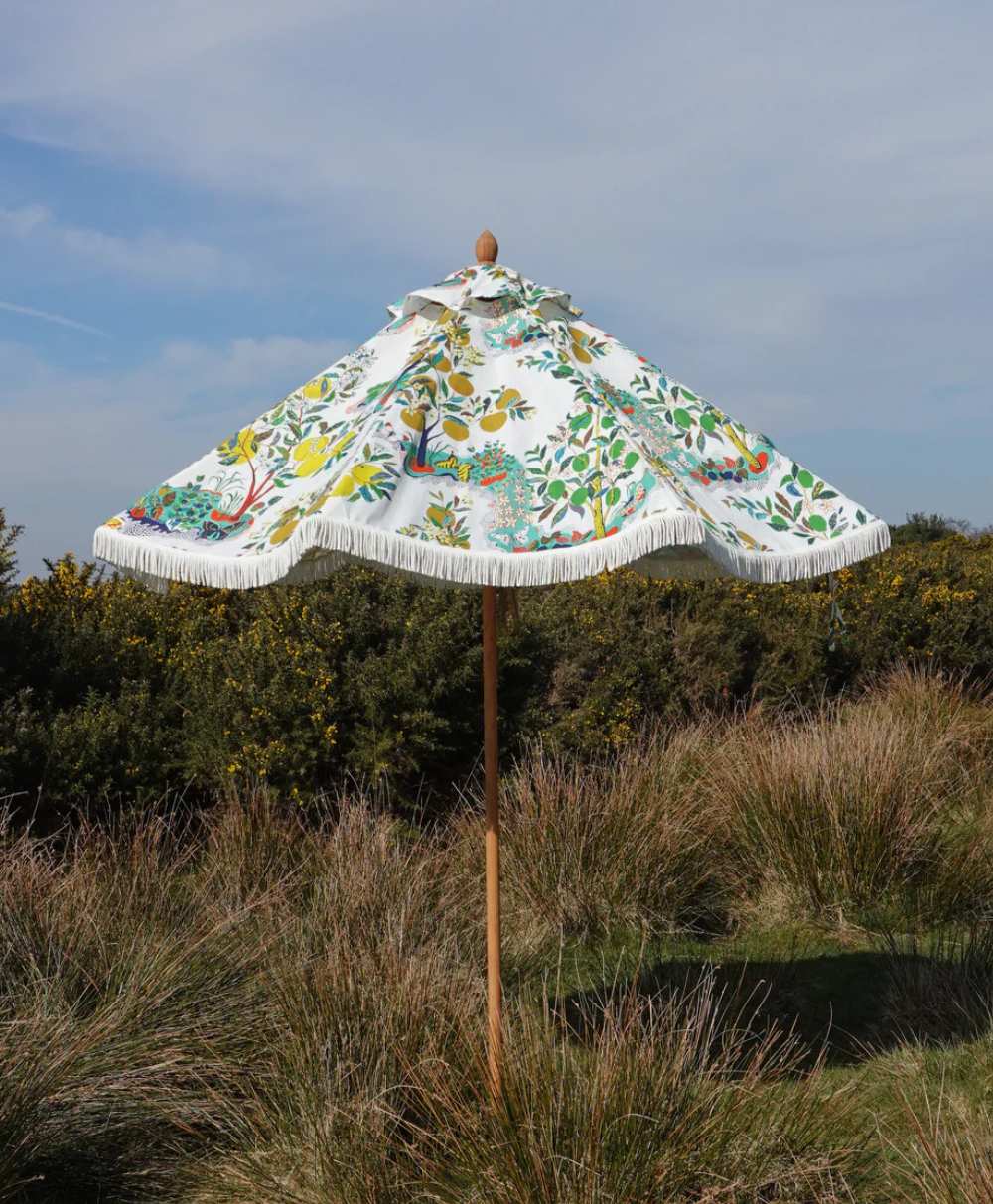 A floral-patterned parasol with a fringe trim stands in tall grass and shrubs under a partly cloudy sky.