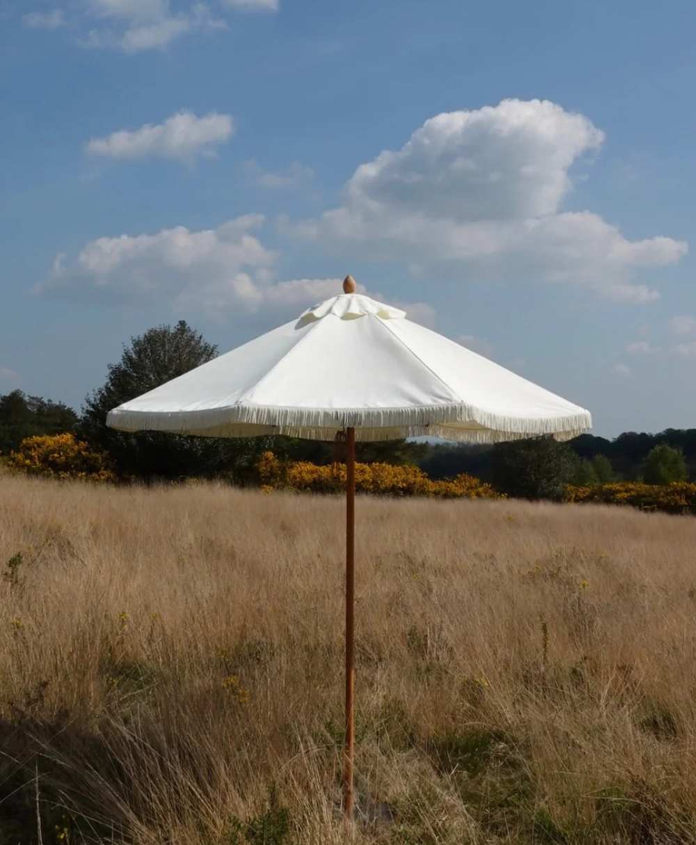 A white patio umbrella with fringe stands in the middle of a tall, dry grassy field under a blue sky with scattered clouds. Trees and bushes are visible in the background.