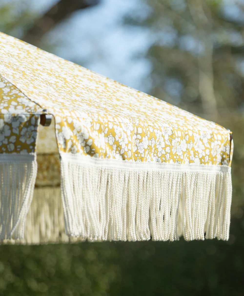 Close-up of a yellow and white floral-patterned outdoor umbrella with long, white fringe trim, shown against a blurred green natural background.