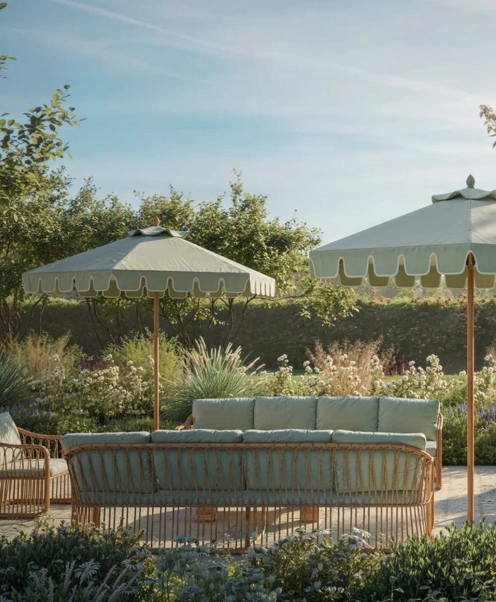 Wicker outdoor sofas with light blue cushions are arranged under two large, pale green umbrellas in a lush, sunlit garden surrounded by greenery and flowering plants.