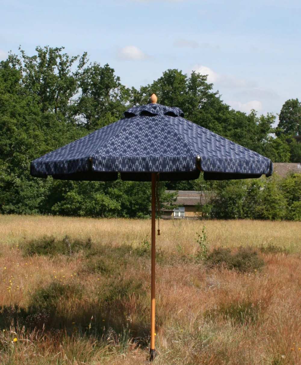 A dark blue patterned outdoor umbrella with a wooden pole stands in a grassy field, with trees and a building in the background under a partly cloudy sky.