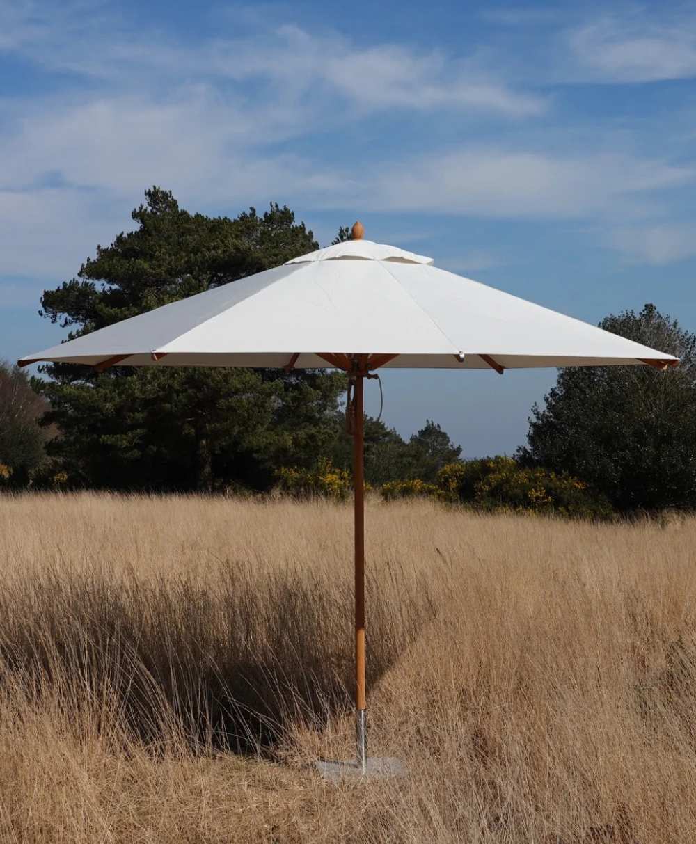 A white patio umbrella stands in the middle of tall, dry grass with trees and a partly cloudy blue sky in the background.