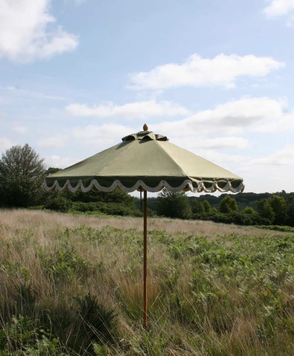 A green patio umbrella with a scalloped edge stands alone in a grassy field under a partly cloudy sky, with trees and hills in the distant background.