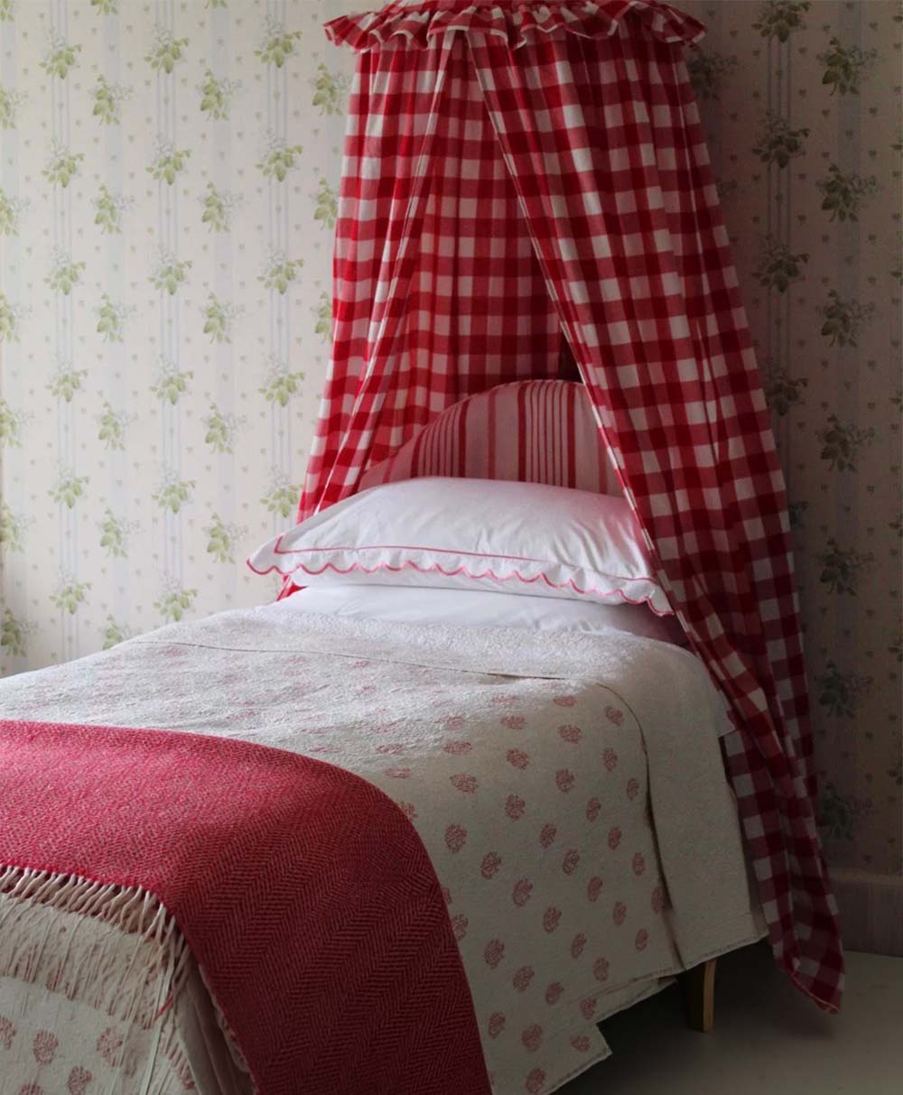 A single bed with white and red bedding, featuring a red and white checkered canopy and matching pillows, stands against a floral wallpapered wall. The room has a cozy, vintage feel.