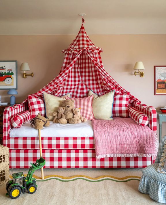 A cozy children’s room with a red and white checkered daybed and canopy, stuffed teddy bears, pastel pillows, a pink quilt, and a toy tractor on the floor. Soft lighting and playful decor create a warm, inviting space.
