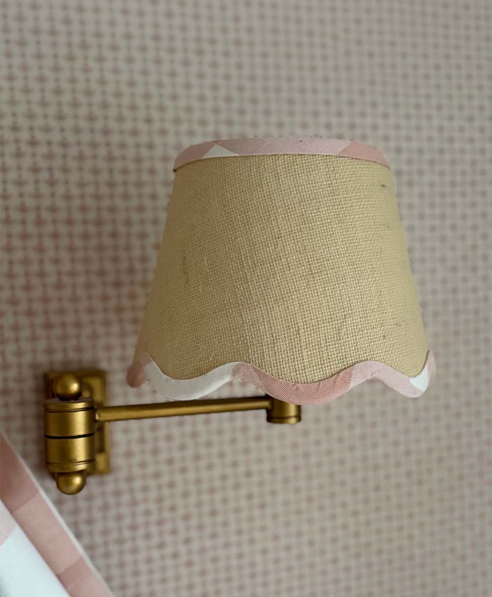 A brass wall-mounted lamp with a beige fabric shade trimmed in pink and white, set against a pink and white patterned wallpaper.