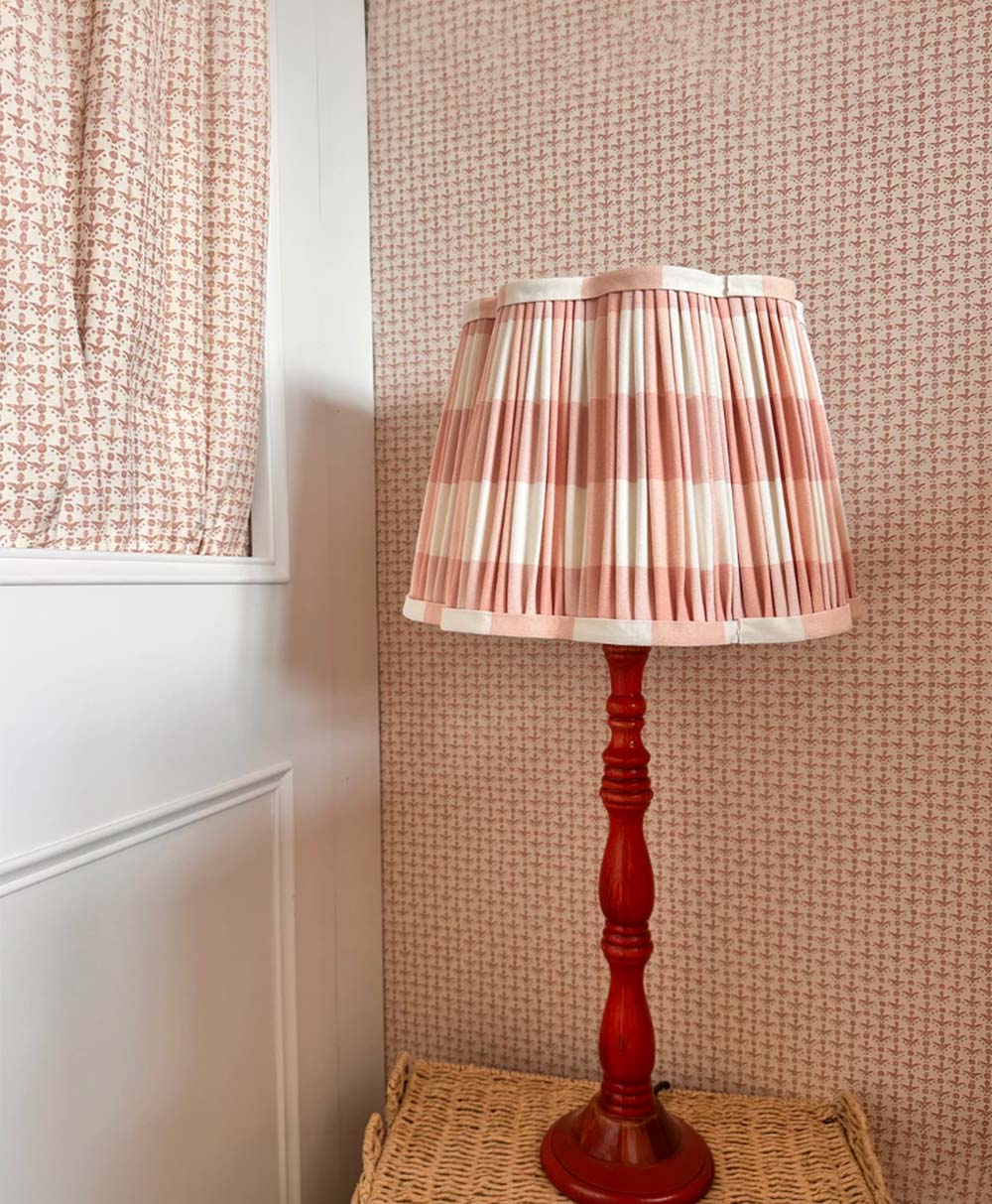 A red table lamp with a pleated pink and white checkered shade sits on a woven surface against a pink patterned wallpaper and near a door with matching pink patterned curtains.