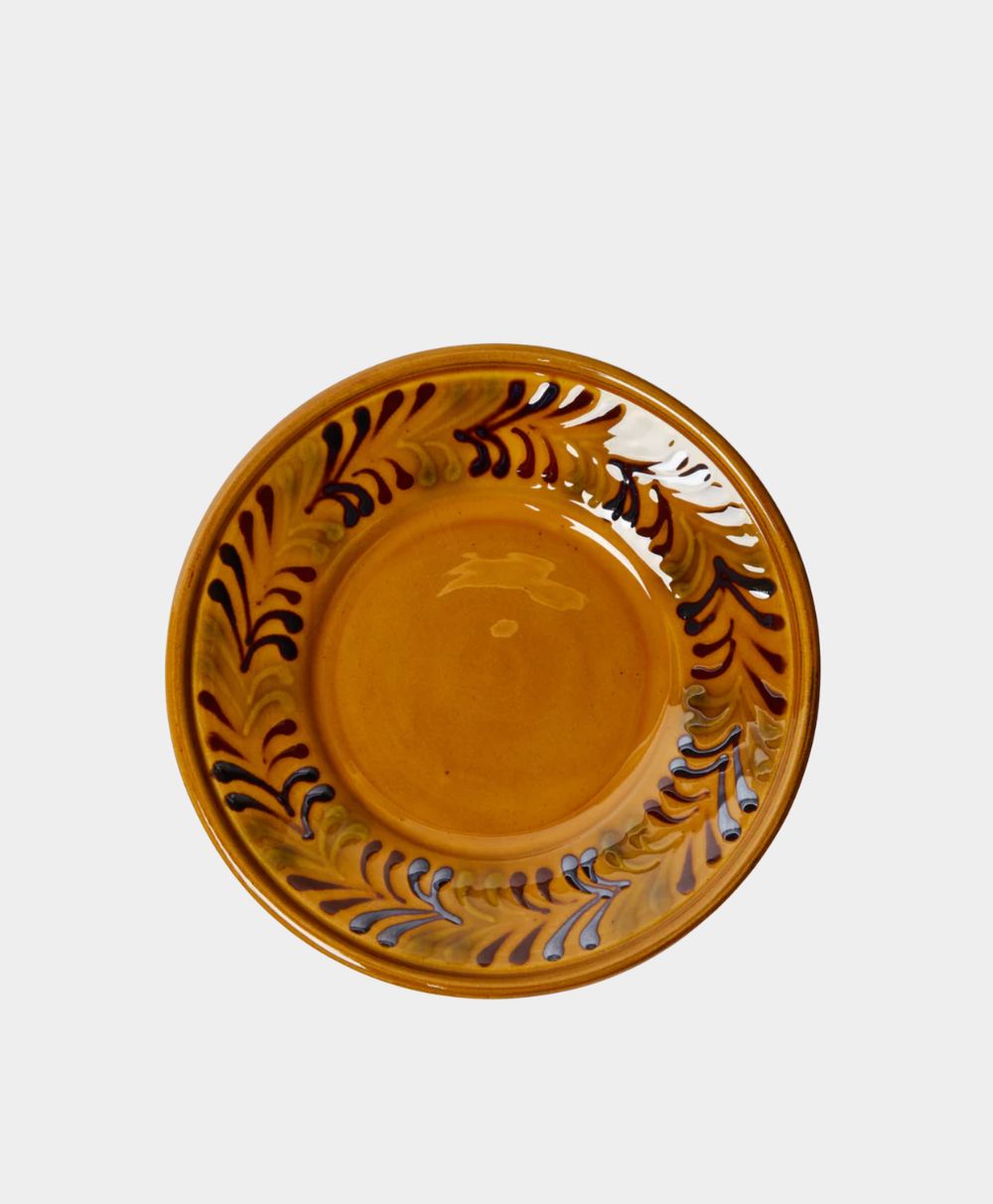 A round, shallow ceramic plate with a glossy ochre glaze, decorated with swirling brown and blue patterns along the rim, set against a plain white background.