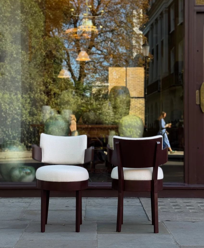 Two modern wooden chairs with white cushions are placed on a sidewalk in front of a glass window, which reflects trees, buildings, and people walking by.