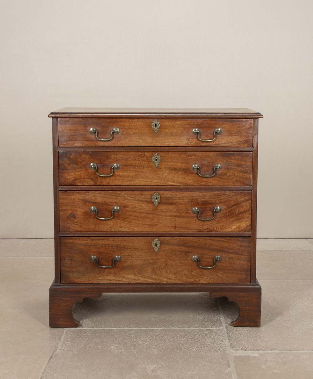 A small antique wooden chest of four drawers with brass handles and keyholes, standing on bracket feet, set against a plain light-colored wall and stone floor.