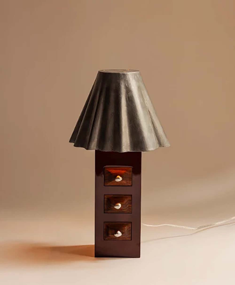 A table lamp with a sculpted, flared metallic lampshade and a rectangular wooden base featuring three small drawers, each with a round white knob, set against a beige background.