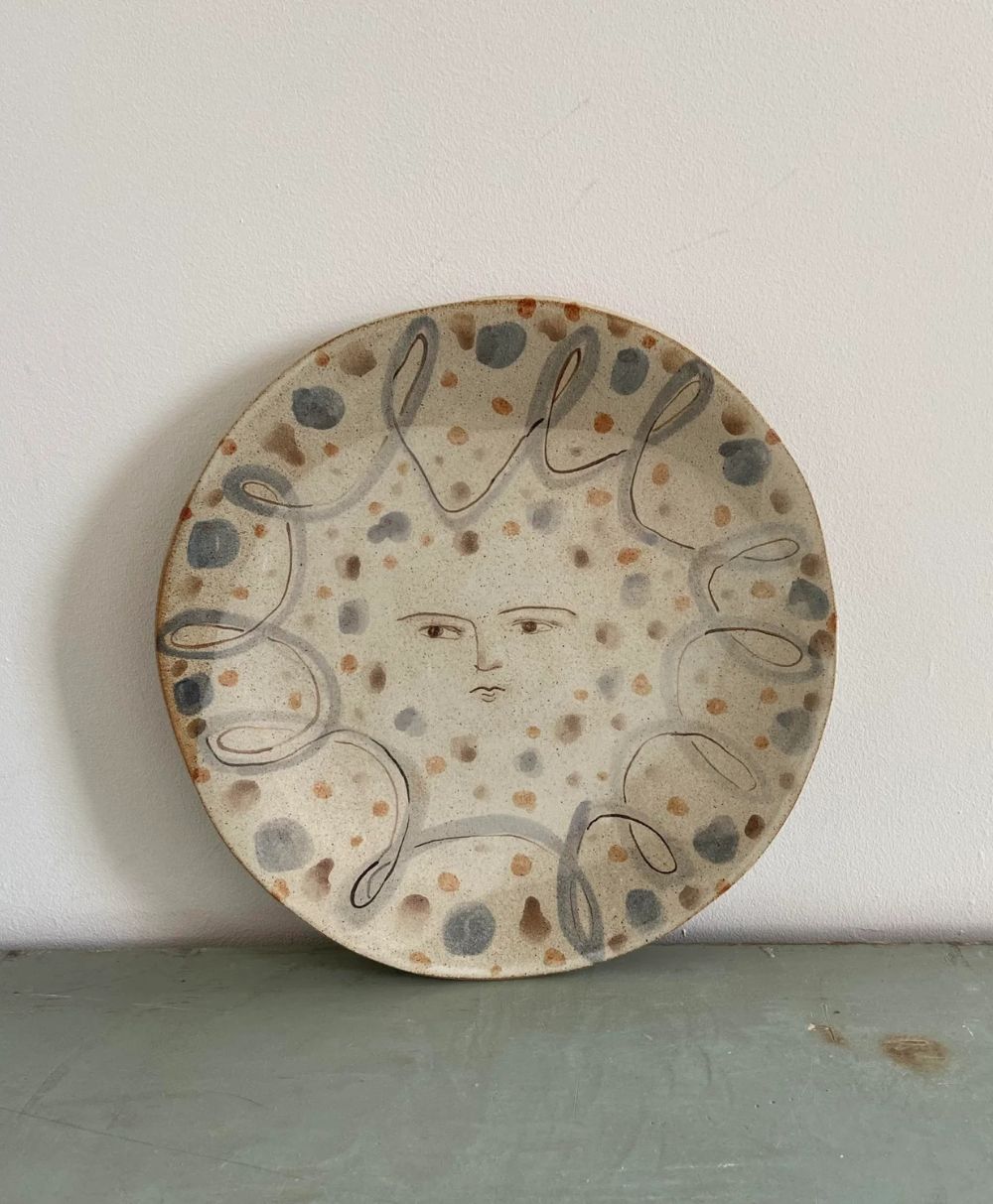 A ceramic plate features a minimalist face in the center, surrounded by abstract swirling lines and scattered gray and brown dots on a light background. The plate leans against a white wall on a gray surface.
